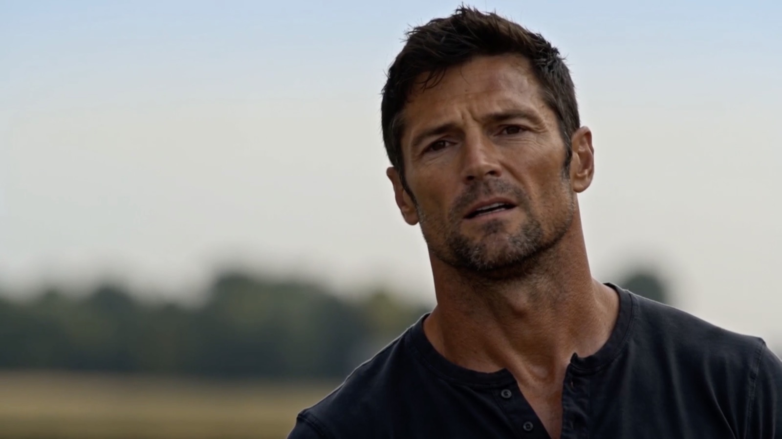 Still from Just Ain’t My Day: a man in a dark henley shirt stands in an open field under a pale sky, caught mid-thought